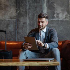 man sitting on a sofa holding a tablet with his suitcase next to him