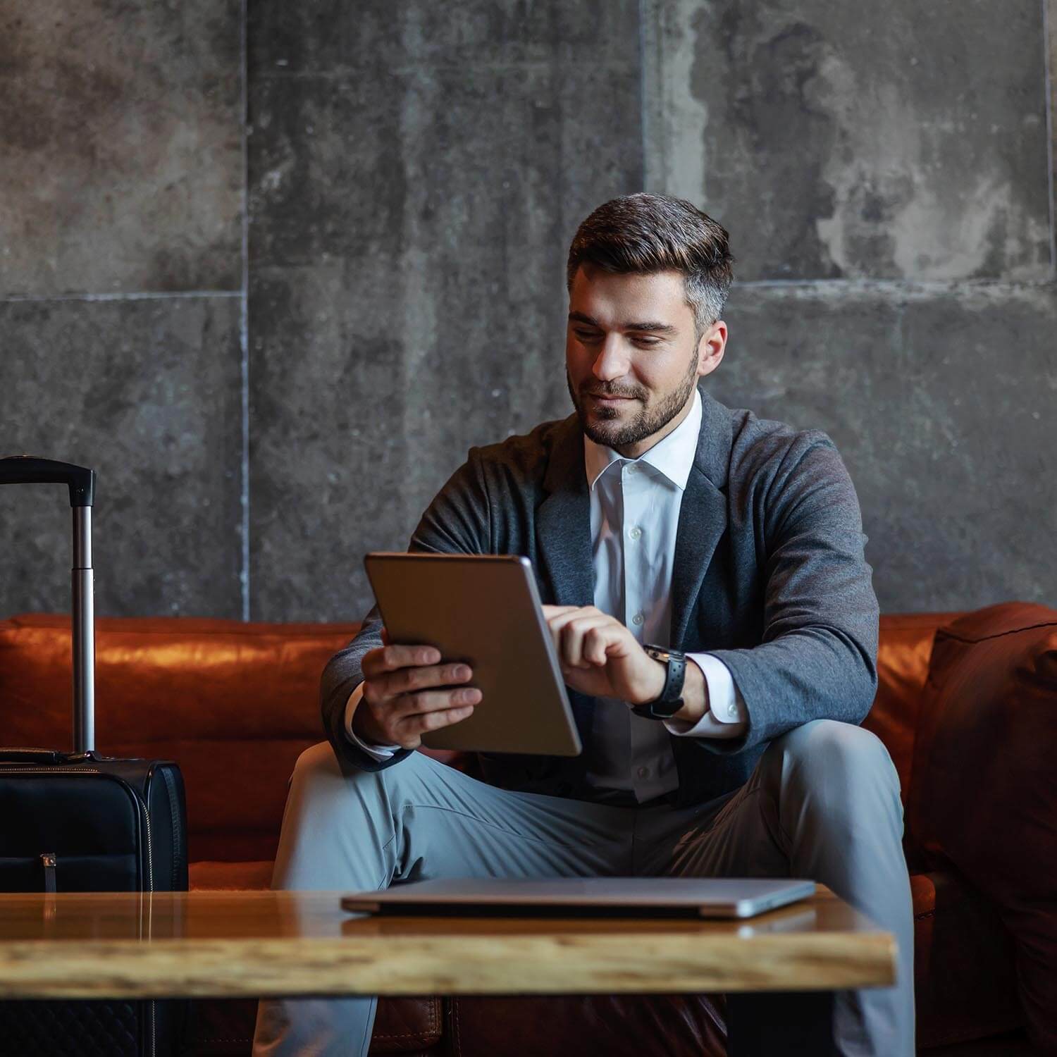 man sitting on a sofa holding a tablet with his suitcase next to him